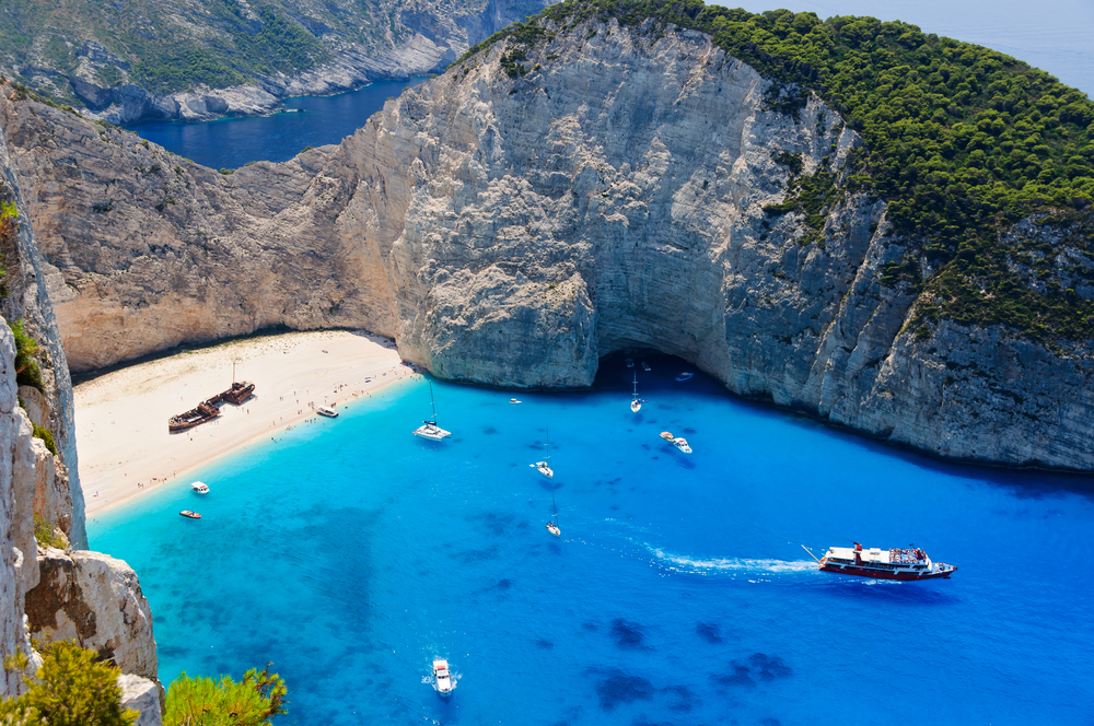 View from the cliffs surrounding Navagio Beach in Greece. The water is a crystal clear blue, the sand is a light tan, there is an abandoned ship on the shore and boats in the water. 