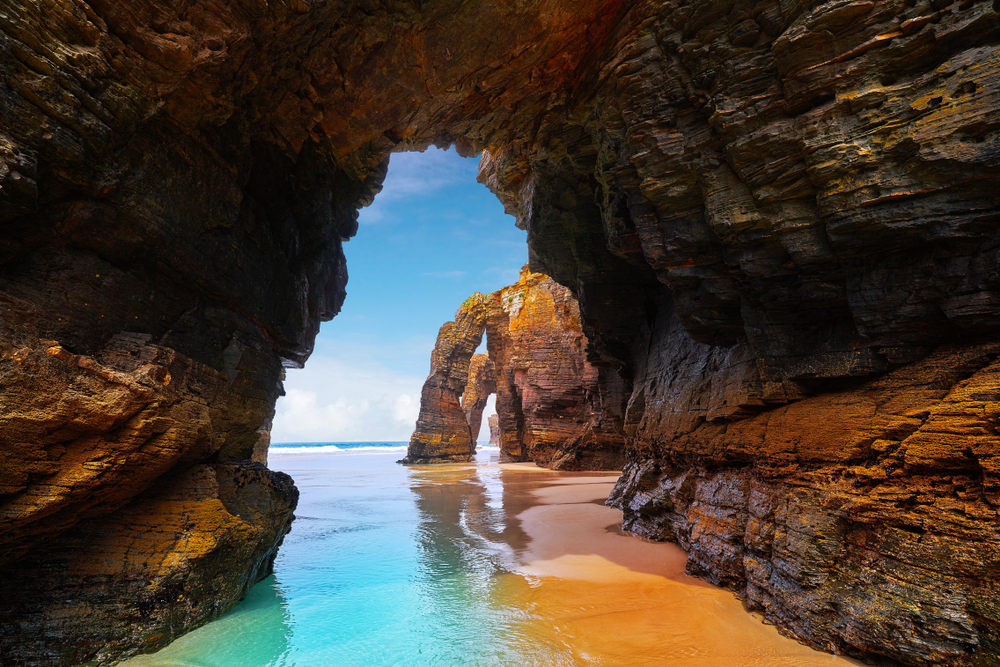 Looking through an orangish red natural stone arch to see blue skies and the turquoise water of Playa de las Catedrales in Spain. 