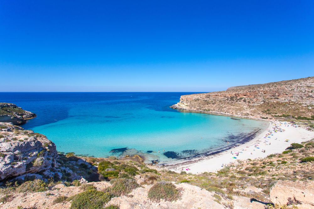 View looking down on Rabbit Beach in Sicily, one of the best beaches in Europe. The beach is in a little inlet, surrounded by a rocky shore. The water is blue and clear. 