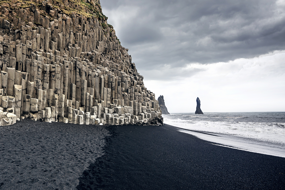 Columnar basalt on the left with a black sand beach and a grey ocean. Reynisfjara Beach is located in Iceland.
