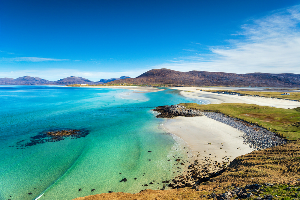 Overlooking Seilebost Beach in Scotland, the sand is white and the water goes from a beautiful green to cerulean. The blue sky is only interupted by low hills in the distance. 