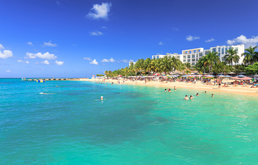 View from the azure water at Doctor's Cave Beach. There are people swimming in the water, umbrellas and palm trees on the beach, and a resort in the background. 