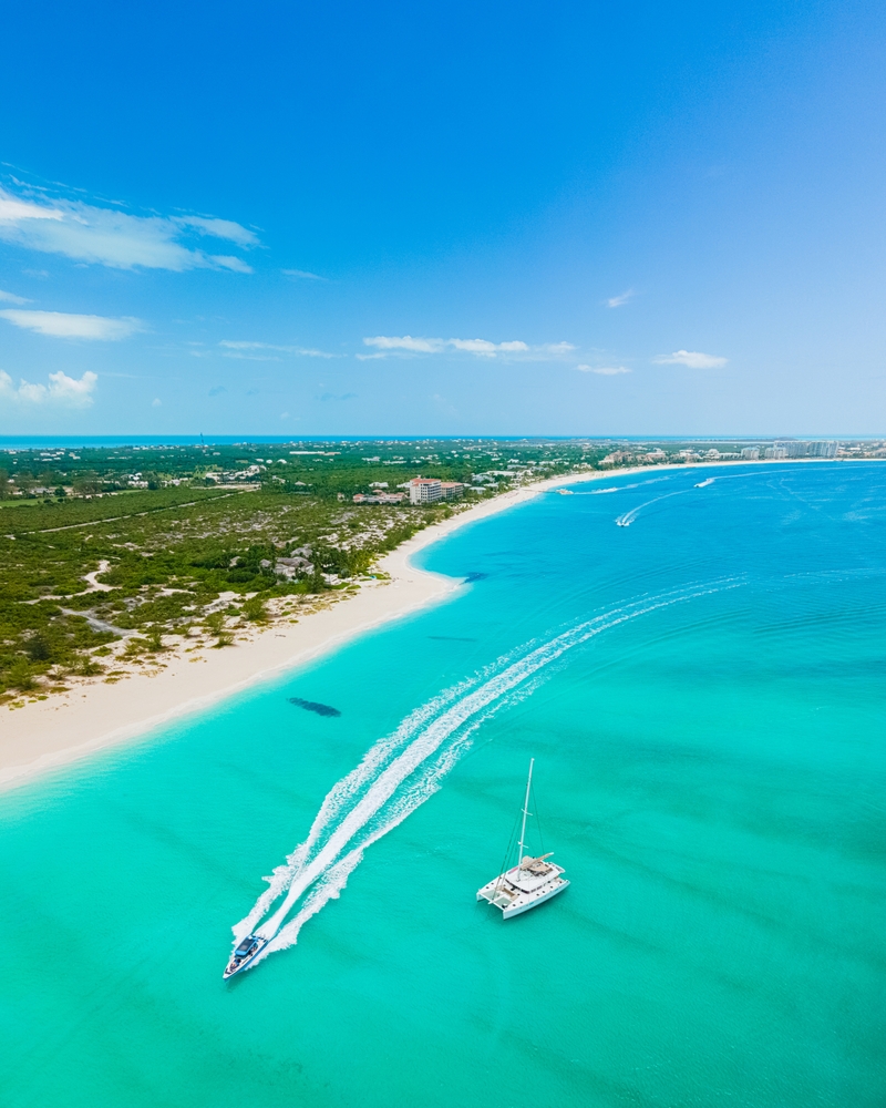 Aerial view of Grace Bay shows a wake boat in the beautiful turquoise water, a strip of white sand, and blue skies. 