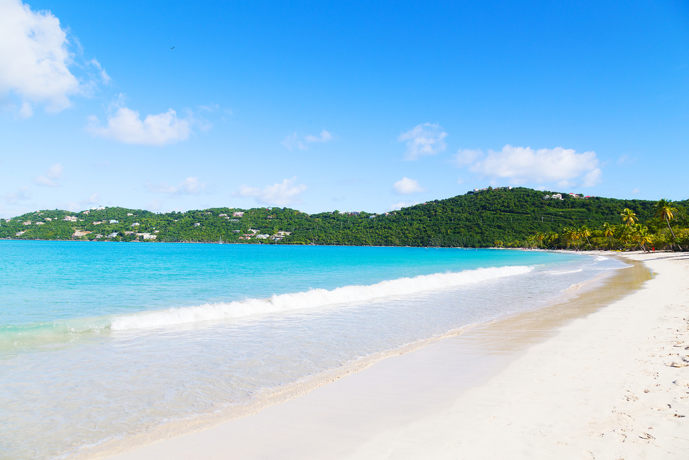 White sandy coastline along bright turquoise water surrounded by a green hilly Magens Bay. 