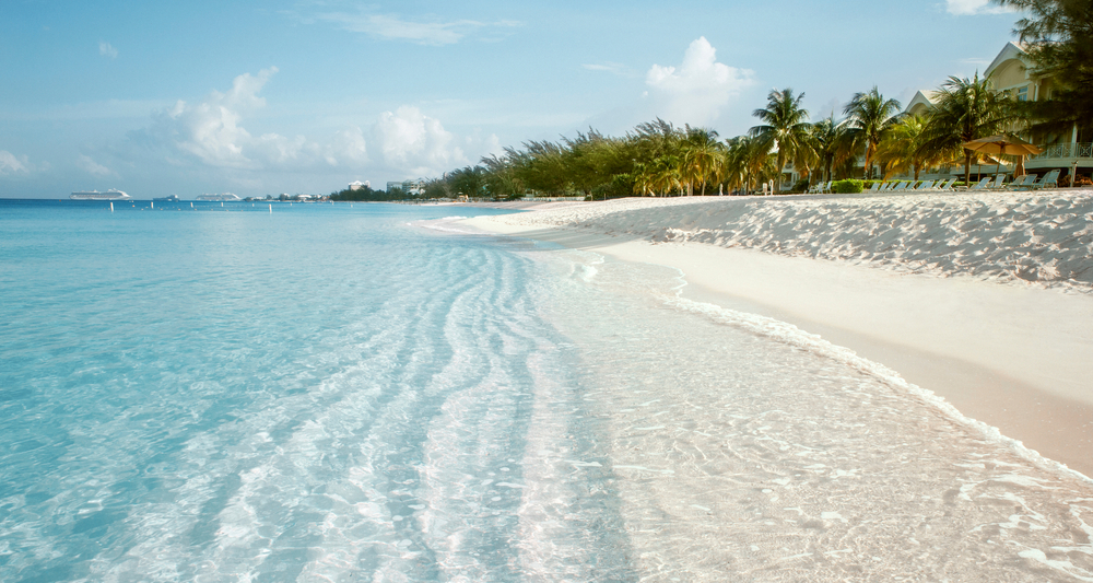 View from just above water level at Seven Mile Beach in Grand Cayman. The water is a crystal clear light blue and the white sand is pillowy soft. There are palm trees lining the beach. 