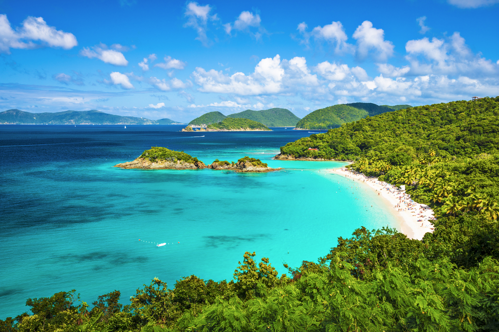 View looking down on Trunk Bay in the US Virgin Islands. The bay is surrounded by a strip of white sand and lots of lush green trees. The water is clear and turquoise. 