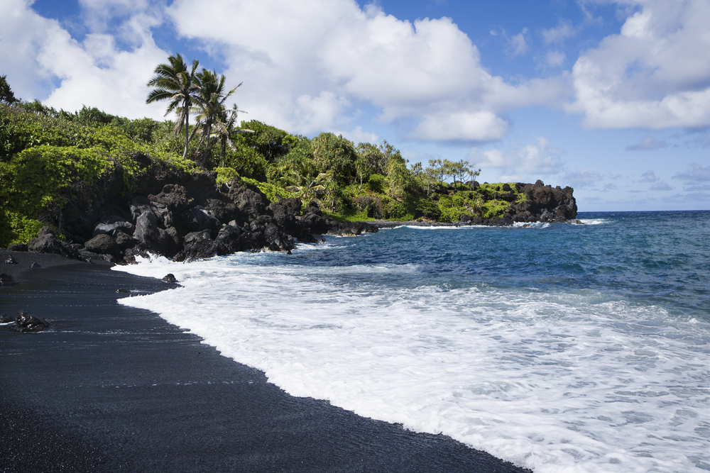 The black sand beach of Honokalani Beach in Maui with an ocean wave receding and green trees surrounding the beach.