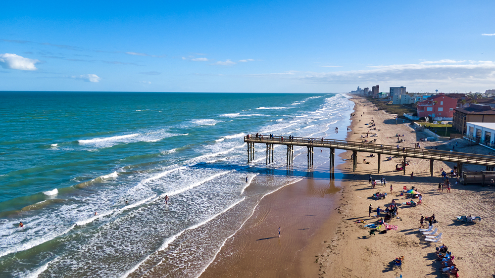 The pier at South Padre Island Beach in Texas extending out into the water. The water is blue green with rolling waves and many people are enjoying the beach. 