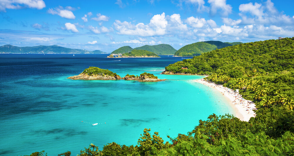 View looking down to Trunk Bay with the green trees on the right, a white sand beach, and vibrant blue water.