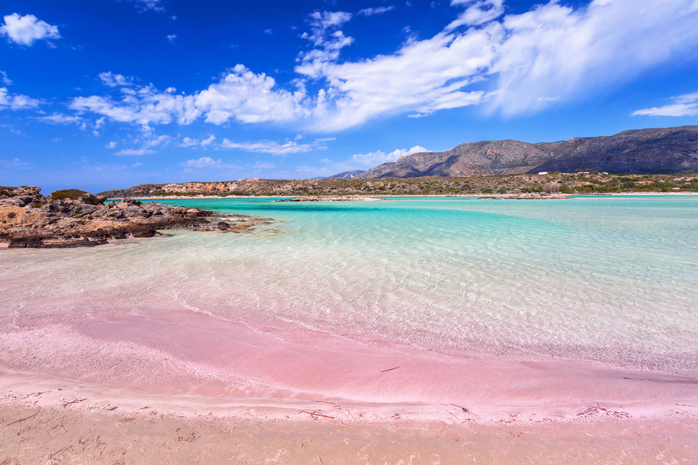 a pink sand beach in Greece where there is clear water and mountains in the background, some clouds are in the sky 