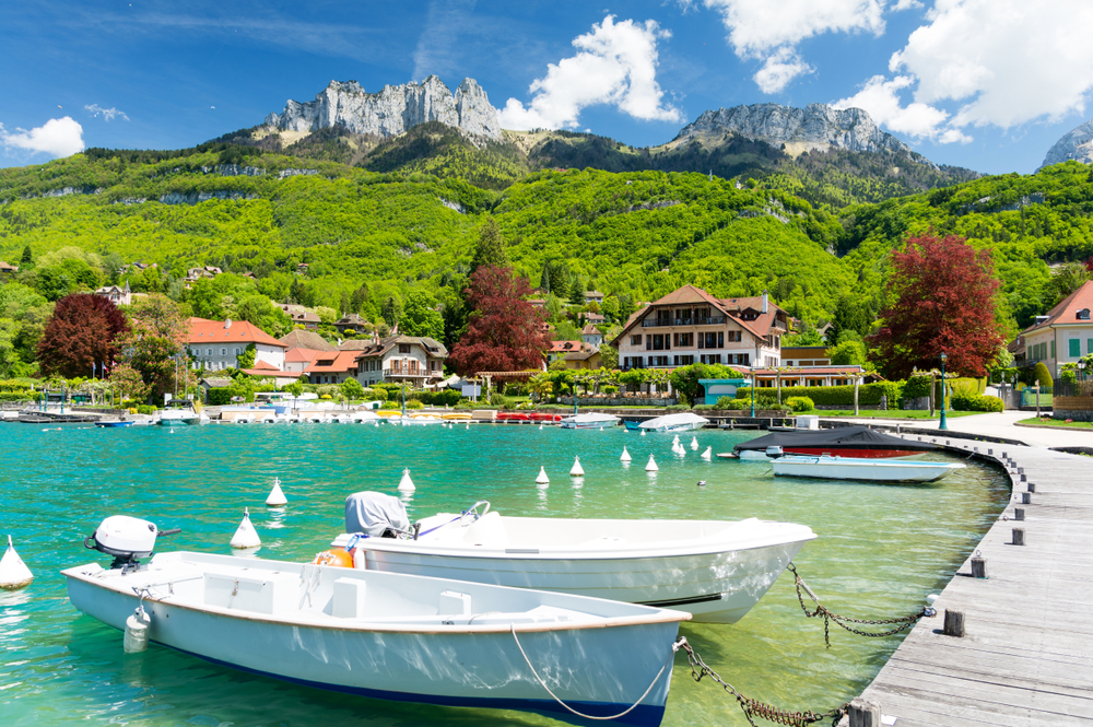 some of the clearest water in Europe, lake annoy, there is a wooden dock on the right, boats docked in the water, and mountains in the background on a partly cloudy day 