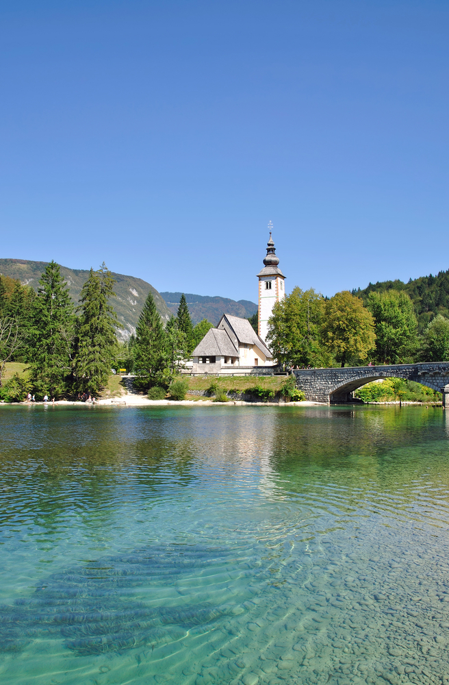 a clear lake with a beach in the background, there is also a church behind the lake and a bridge on the right 