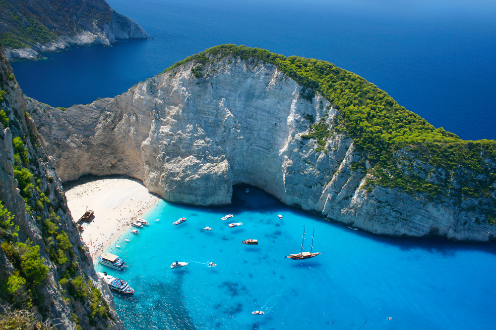 aerial photo of ocean, white sandy beach, and rock cliffs with trees on top, there is a shipwreck on the beach and there are boats docked in the water 