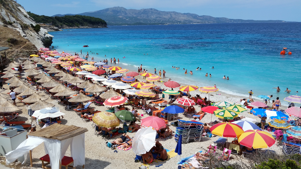 a beach in Albania, there are many umbrellas and sunbeams laying out on the beach, there are people swimming in the water 