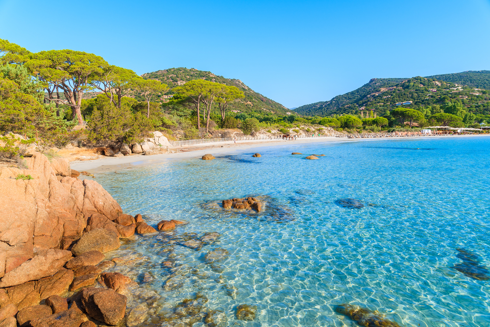 mountains in the background, a sandy beach in the middle of the photo, water is taking up the rest of the photo, there are some rocks in the water, the sky is clear 