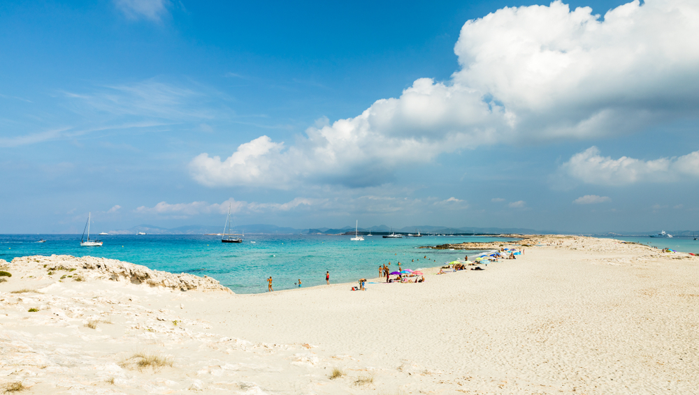 one of the best clear water beaches in Europe, a slender sandy beach with crazy clear/blue water on both sides, people are laying out on the beach and there are boats in the water 