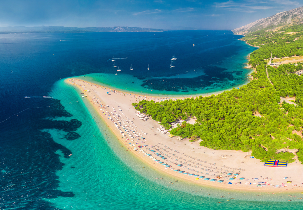 aerial photo of a Long Beach with clear water, there are trees on the right and mountains in the background 