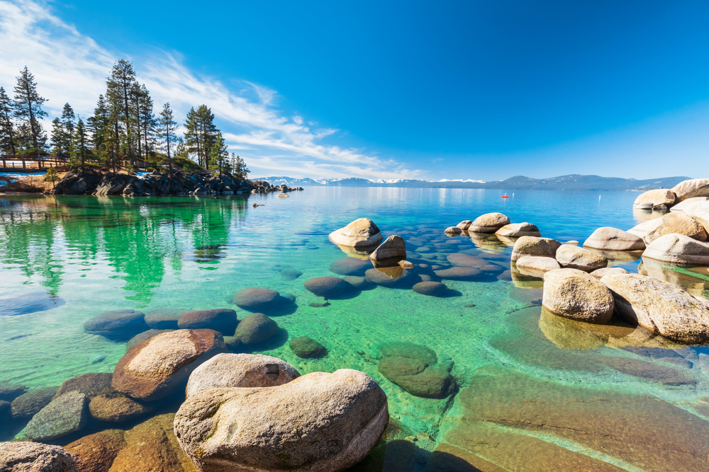 Rocks and stones line the bottom of this crystal clear lake, and trees and mountains crowd the shoreline in the background of the photo. 