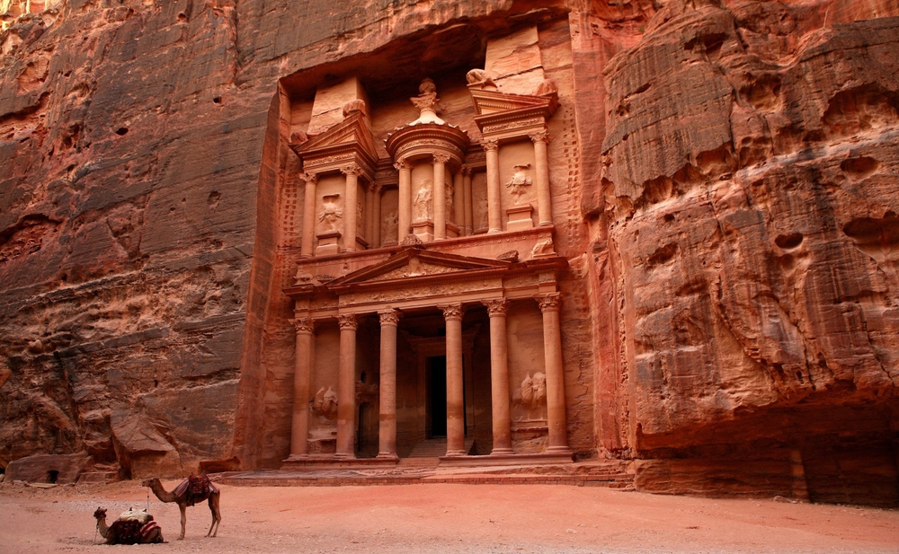 Petra Jordan. brown concrete building, carved into the rock. You can see two camels in the foreground. 