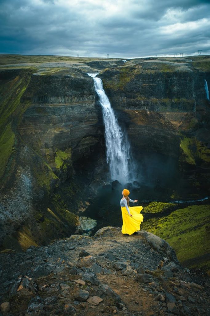 Girl standing on a rock arms out spread with a waterfall in the background. She is wearing a yellow skirt.   
