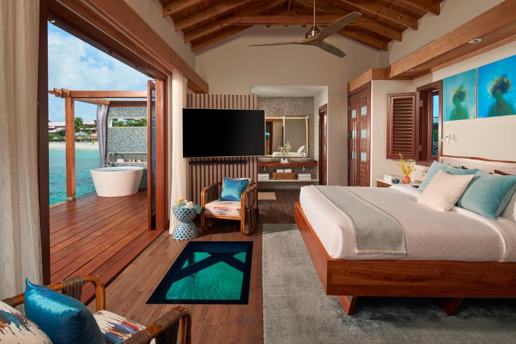 Inside of a water villa showing a bed a chair and opine doors leading to the deck with a bath on it. You can see water in the background.  