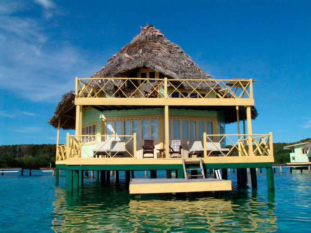 Close up of a water villa that has two stories and a large deck. There are chairs on the deck. The villa is green with a thatched roof. 