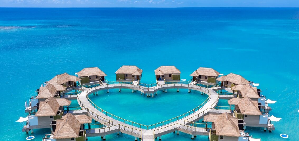 drone shot of heart shaped overwater bungalows with two people walking out to them over blue water