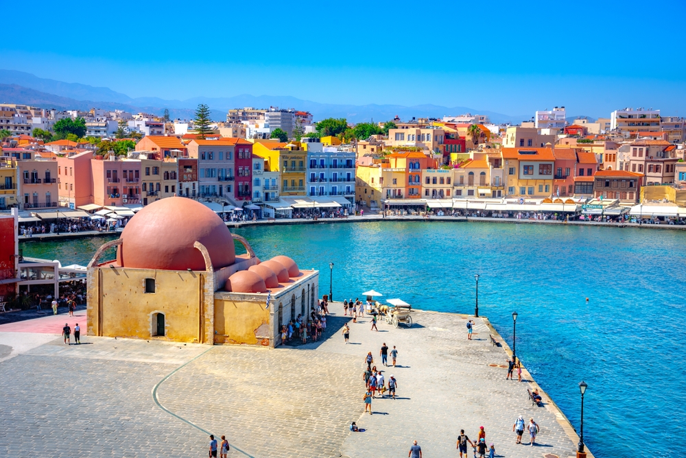 a domed building on the waters edge, in the background there are colorful buildings along the edge of the water on a clear day, people are walking along the waters edge