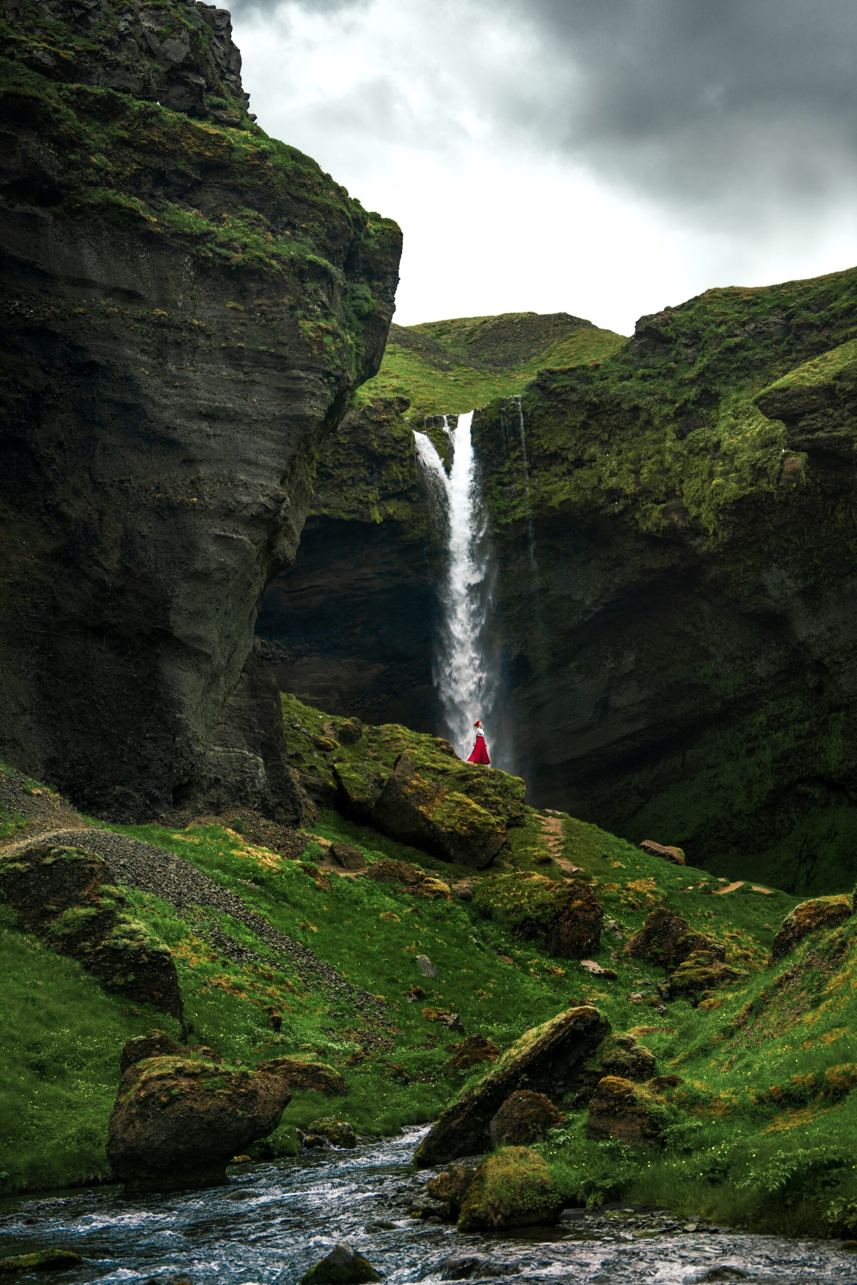 a waterfall is falling down from a cliff above where a woman is standing, she is wearing a long red skirt 