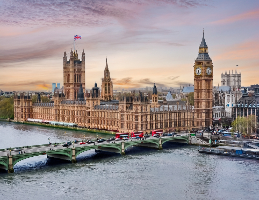 a bridge is crossing a river in London England, buildings are in the background including Big Ben, a UK flag is flying high above the buildings 