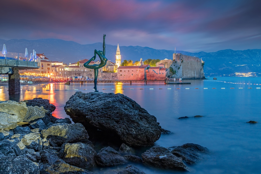 there is a statue of a woman doing a yoga pose on a rock on the edge of the water, a city with lights on is in the background, and behind that there are mountains