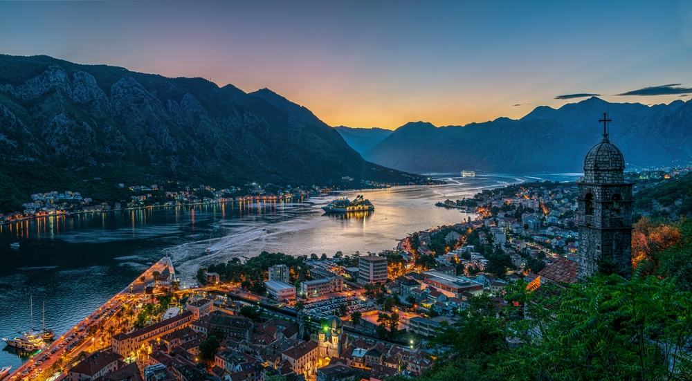 water in between two mountains, city lights and buildings are on both sides of the water, a boat is going through the middle of the picture 