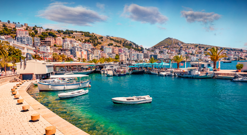 places to visit in Europe in August, the Albanian coast on a partly cloudy day, boats are in the water, palm trees are on the edge of the water, buildings scale the mountains in the background