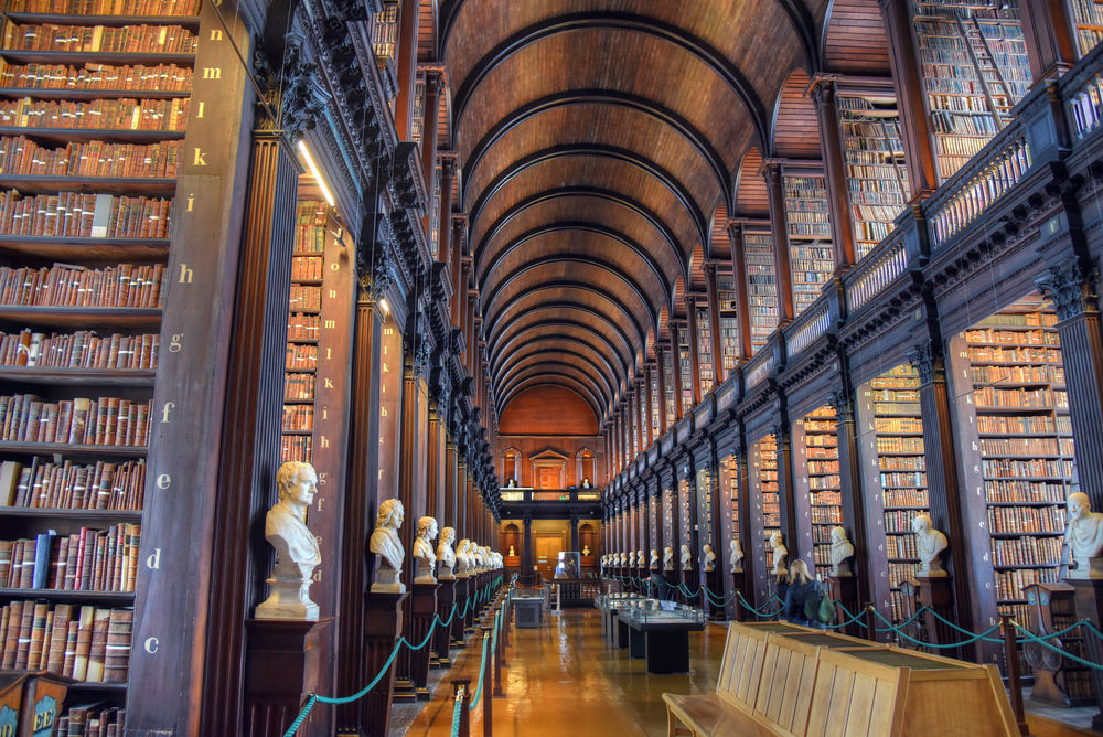 View down the Long Hall in Trinity College in Dublin with a high, curved ceiling and two stories of bookshelves and busts of people.