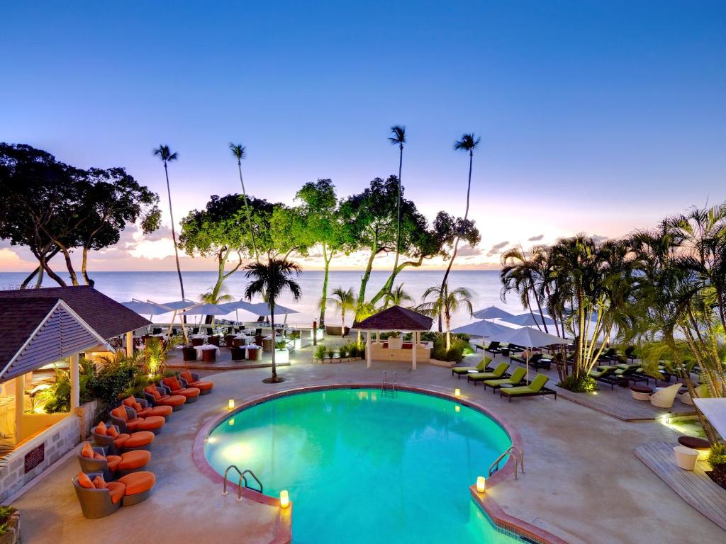 The pool at dusk is lit up, surrounded by candles, loungers, umbrellas and palm trees. The ocean is purple in the background. 