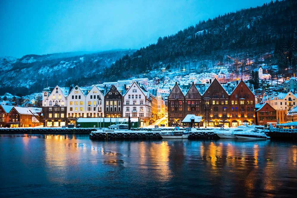 water in the foreground, buildings line the edge of the water in the centre of the image and there are tree covered mountains in the background, one of the best places to visit in europe in december 