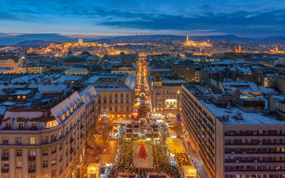 a town square with a christmas tree and lights in it as well as tents/stall in a Christmas market, the city stretches out into the back of the image and there are mountains in the background, one of the best places to visit in europe in december