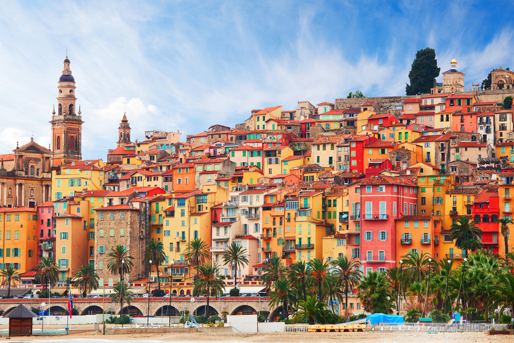 a hillside village of colorful buildings, there is a tower on the left, palm trees in the foreground and partly cloudy sky, french riviera, one of the best place to visit in europe in december 