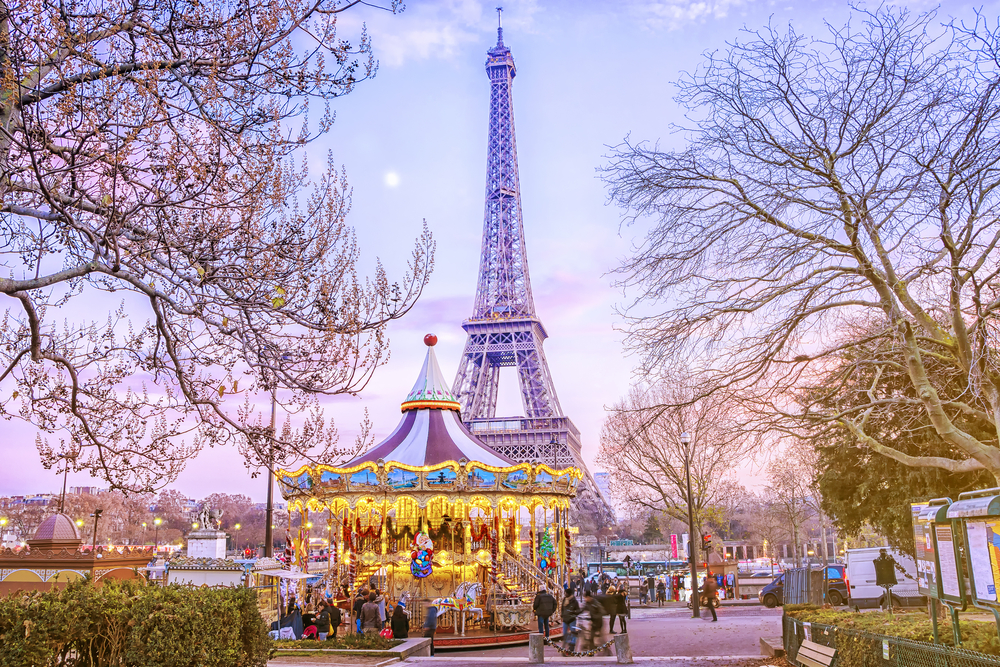 Eiffel tower in the middle of the photo in the background, a carousel is in front of it and there are trees om both sides of the image 