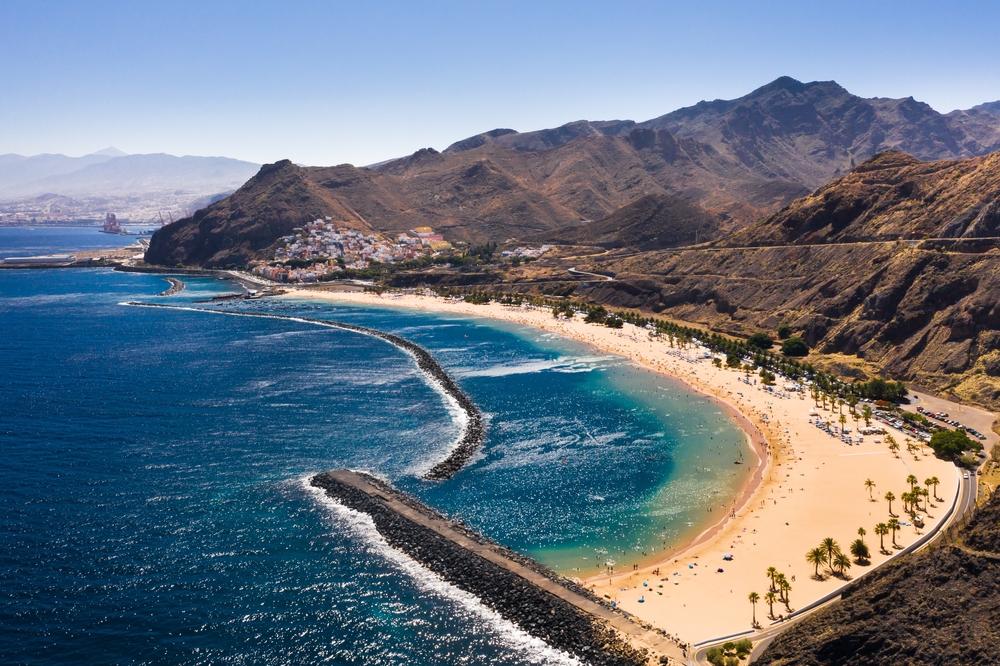 sandy beach on the coast, water on the left, mountains on the right, the beach is curved and there are palm trees on it 