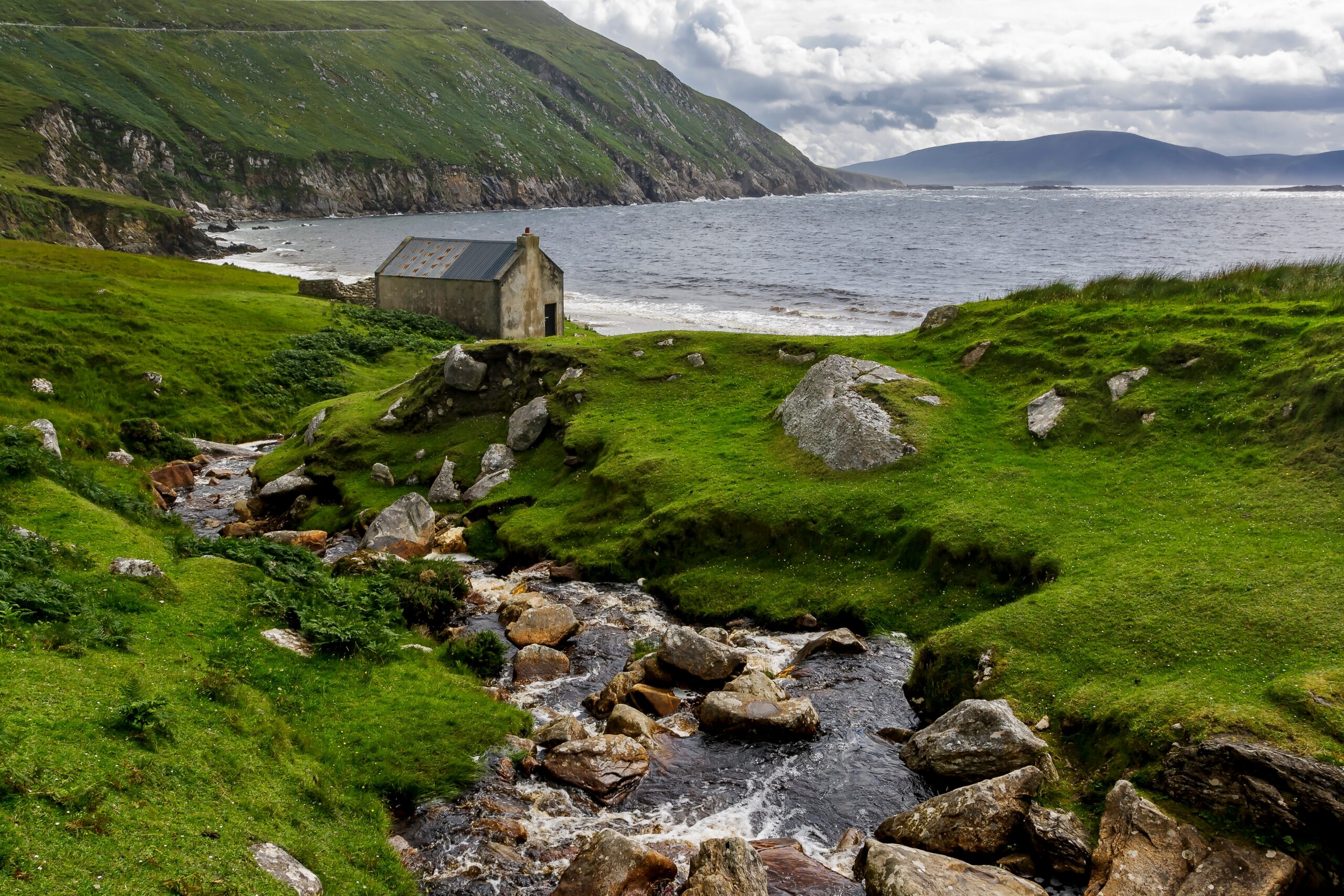A creek running to a dramatic beach surrounded by hills and other large island. The beach has a classic Irish stone cottage on it. 