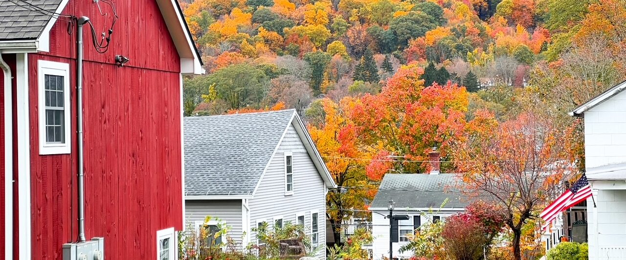 red barn with white trim to the left of the photo with white houses in the background and fall foliage up on the side of the mountain