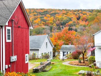 red barn with white trim to the left of the photo with white houses in the background and fall foliage up on the side of the mountain