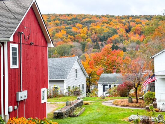 red barn with white trim to the left of the photo with white houses in the background and fall foliage up on the side of the mountain