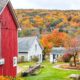 red barn with white trim to the left of the photo with white houses in the background and fall foliage up on the side of the mountain