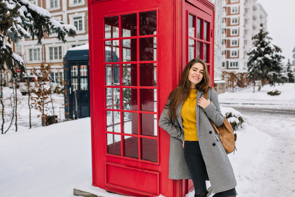 Woman in trendy yellow sweater posing with pleasure next to red phone booth in winter. One of the London outfits in January.