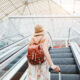 woman standing on escalator wearing hat and brown backpack at the airport in summer