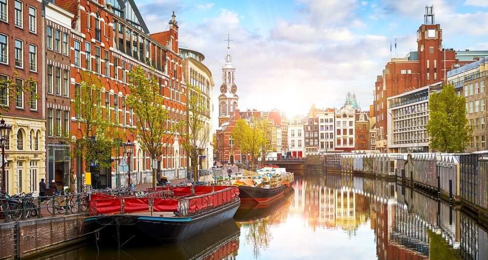 The canal city of Amsterdam with stunning architecture and bridges going over the canal. the biggest boat is black with red interior. the sky is blue with some white clouds and there are brown and reddish color buildings on either side of the canal.