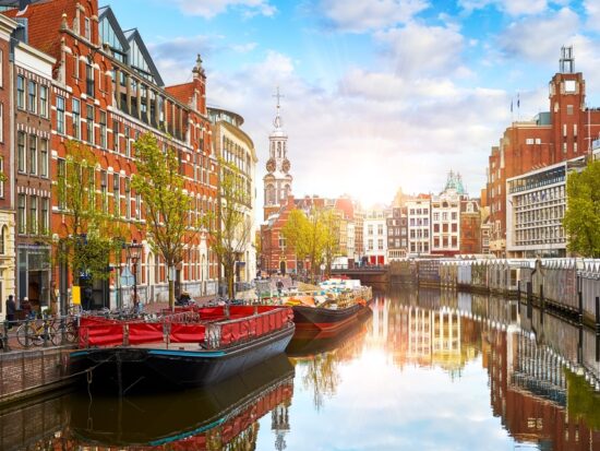 The canal city of Amsterdam with stunning architecture and bridges going over the canal. the biggest boat is black with red interior. the sky is blue with some white clouds and there are brown and reddish color buildings on either side of the canal.
