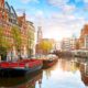 The canal city of Amsterdam with stunning architecture and bridges going over the canal. the biggest boat is black with red interior. the sky is blue with some white clouds and there are brown and reddish color buildings on either side of the canal.